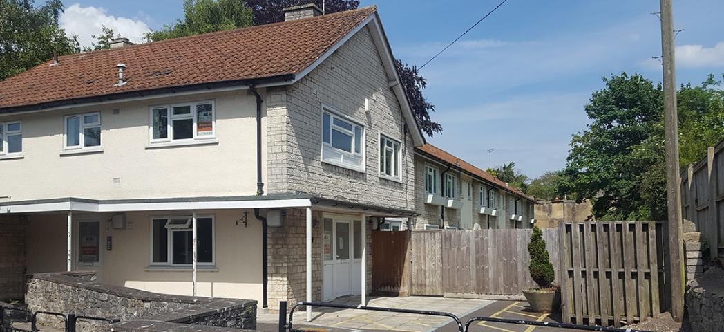 A two-story residential building with a light-colored exterior and a red-tiled roof is visible. In front of the building, there is a gated entrance leading to a parking area. The surrounding area features a stone wall and a wooden fence, with trees and greenery in the background. The sky is clear with a few clouds.