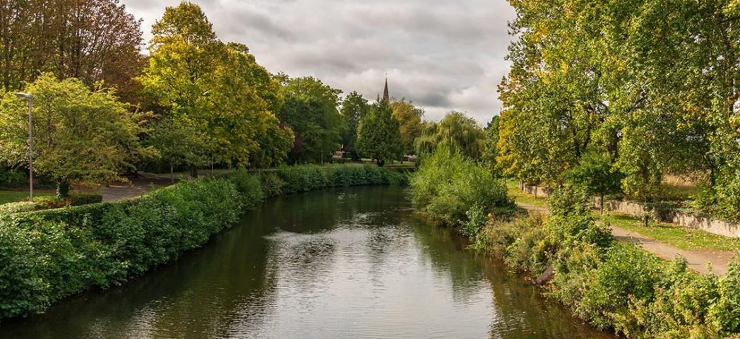 A calm river bordered by lush green trees and shrubs, with a church spire visible in the distance under a cloudy sky