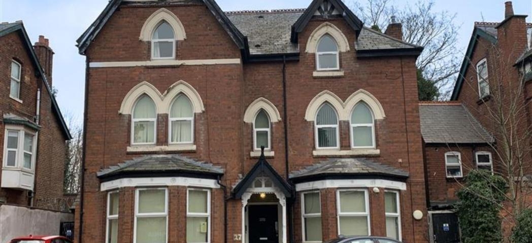 A large, three-story Victorian-style brick house with decorative gables and arched windows. The building features a symmetrical facade with a central entrance and two large bay windows on either side. The roof has a steep pitch, and there are trees in the background. A parked car is visible in front of the house, and the sky is partly cloudy.