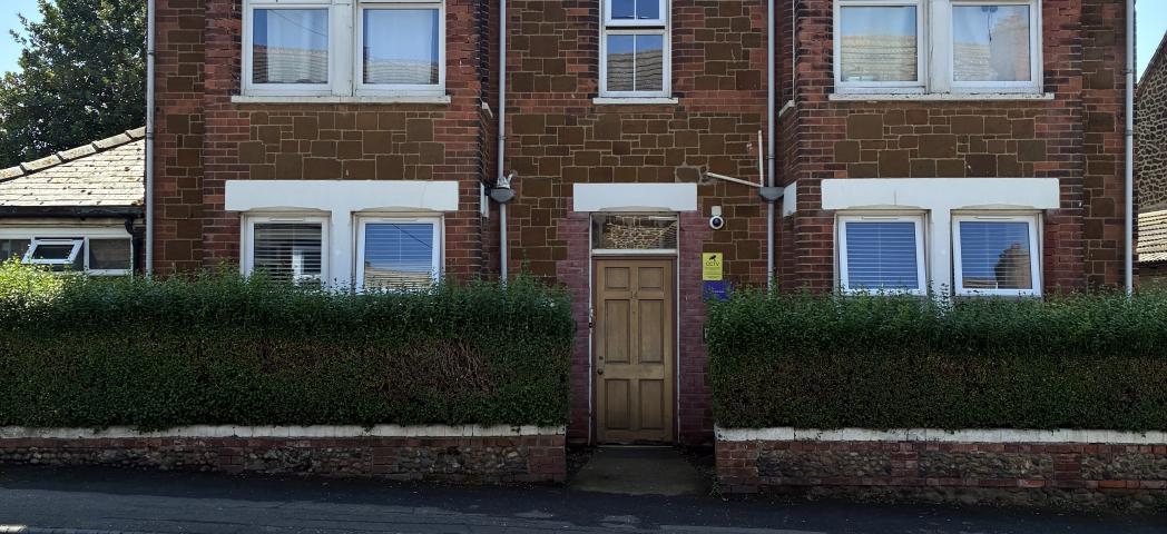 A 3 story dark brown brick building with white window panes. A hedge surrounds the front perimeter with a gap in the middle to allow entry