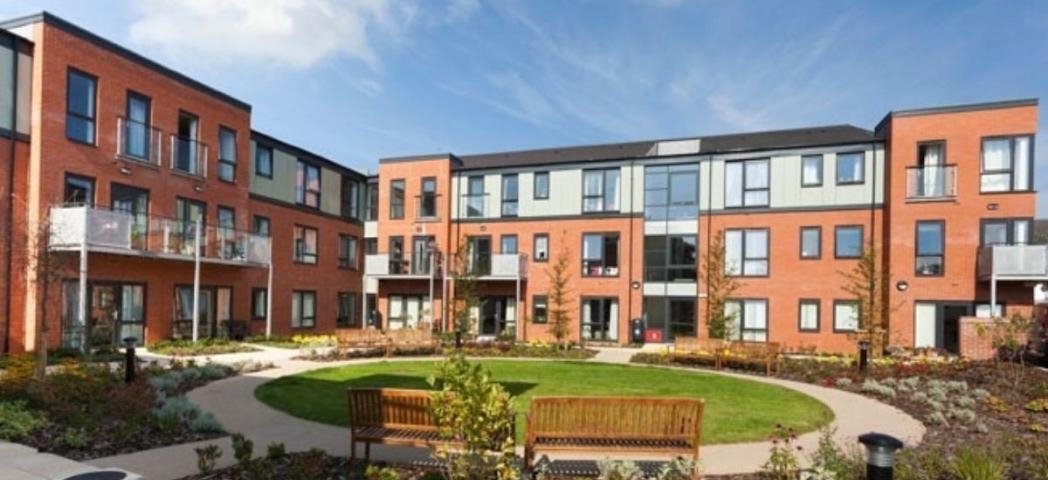 A modern, multi-story brick apartment building with large windows sits under a bright blue sky with scattered white clouds. A circular garden with a well-maintained green lawn and flowerbeds surrounds the building, featuring a wooden bench and pathway.