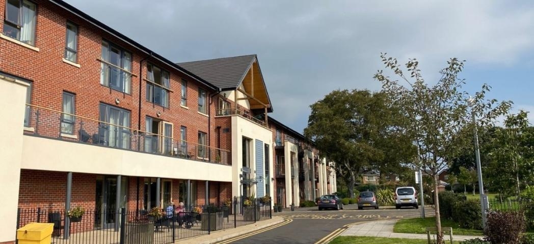 A street scene featuring a row of modern, multi-story brick apartments under a cloudy sky. A paved road curves gently ahead, with several parked cars and a yellow traffic cone visible. Trees and landscaping flank the road, and a small seating area is present at the front of the buildings.