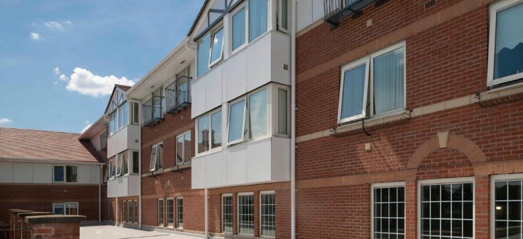 Exterior view of a multi-story brick building under a bright blue sky. The building features several windows with balconies on the upper floors, and a paved courtyard extends to the left. A decorative stone pillar is visible in the foreground.