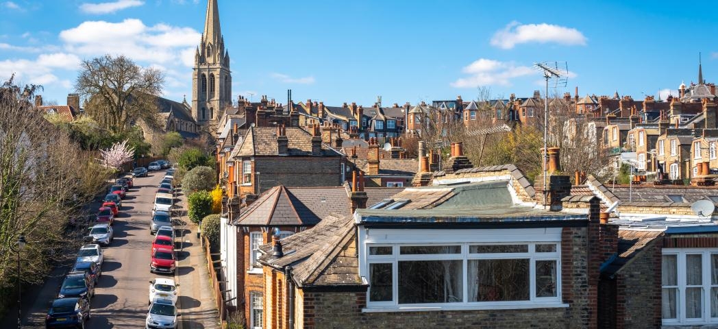 Residential street lined with Victorian houses and a church spire under a blue sky.