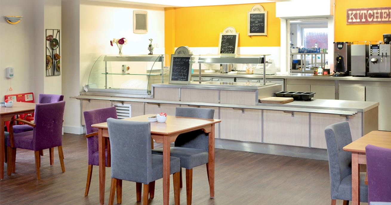 Purple and silver suede chairs tucked under small four person wooden dining tables. These lead to a industrial serving hatch with warming areas and chalk board menu. Behind it are coffee machines and food preparation areas. 