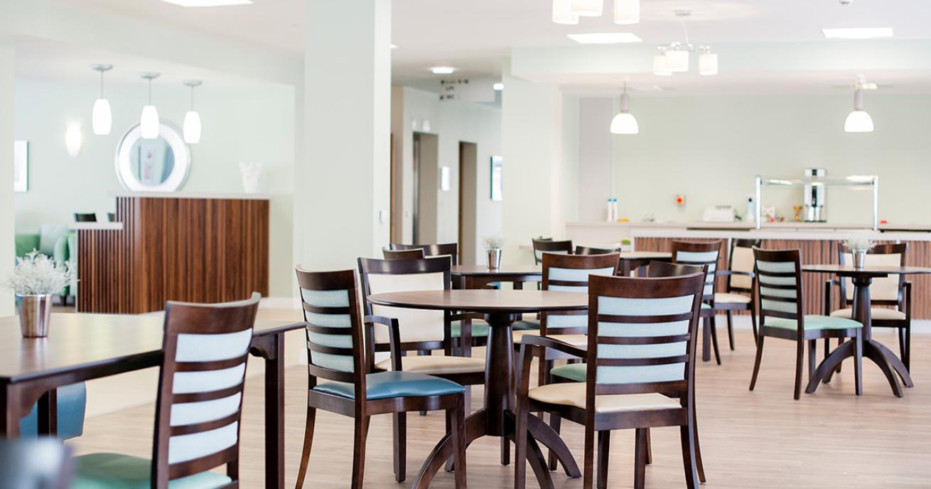 Spacious and clean restaurant area. Muted blue painted walls with multiple hanging light fixtures a large wooden serving area with industrial heat lamps. A vast selection of mahogany circular dining tables and chairs with duck egg blue upholstery.