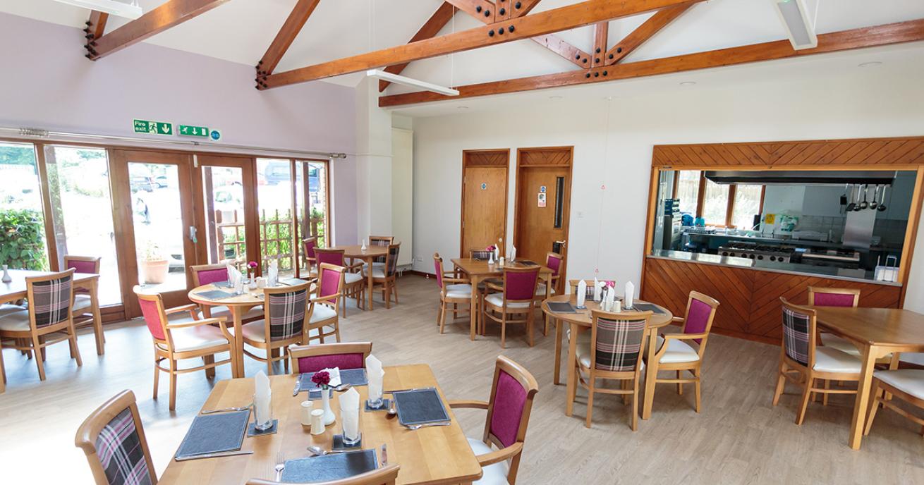 A roomy dining area with access and views into the communal garden An array of four person light wooden dining tables sets are located in front of the restaurant serving hatch. 