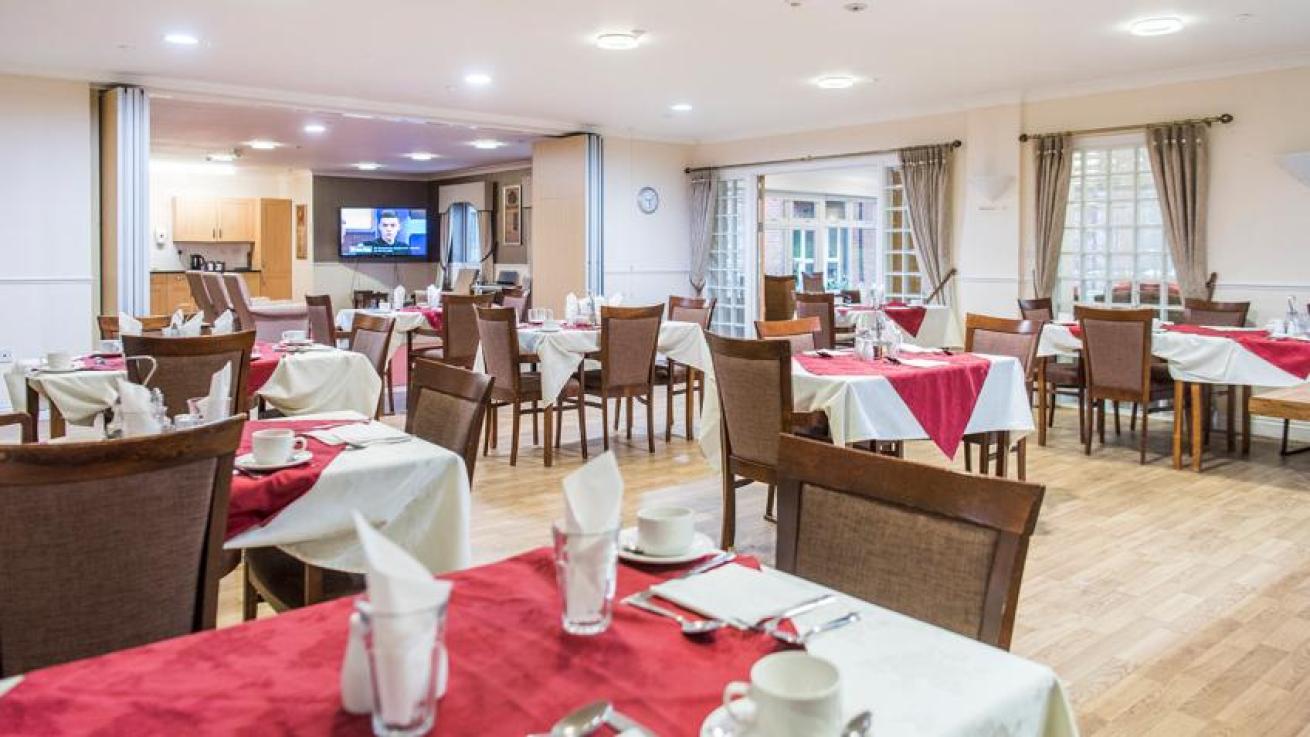 Spacious, light dining area with white and red linen dressed dining tables, set with teacups, saucers, cutlery, and napkins. Set in a floral silver and white patterned decorated room, with matching silver hanging picture frames. As well as silver crushed velvet curtains in front of the large windows and French doors. 