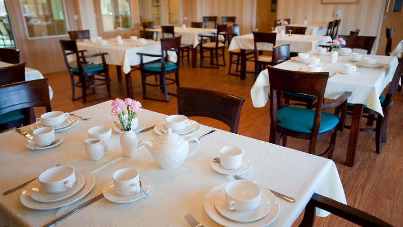 The traditional dining room is home to multiple mahogany dining tables dressed in white linen cloths and matching chairs with blue coloured leather upholstery. The tables are dressed with white ceramic plates, soup bowls and tea set with silver cutlery.