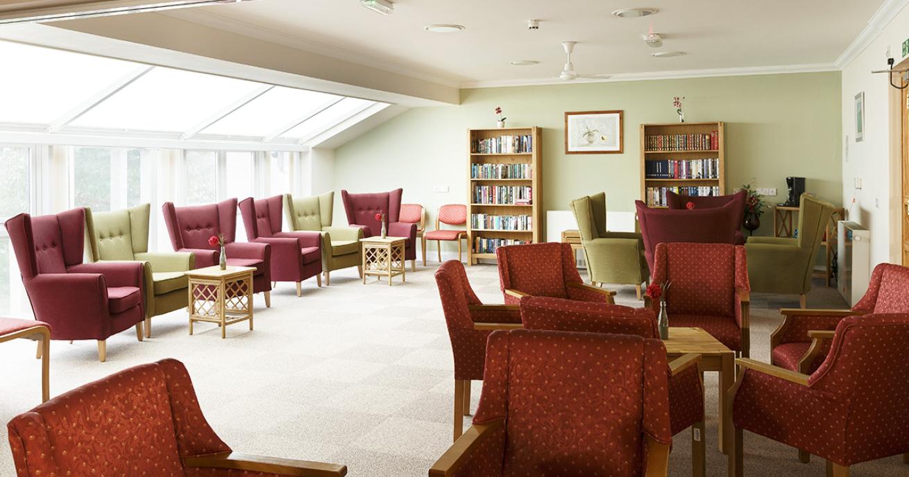 A calming seating area with a collective of burgundy and pea green chesterfield armchairs paired with small woven coffee tables. Two tidy wooden bookshelves sit centrally on the pale green wall. Natural light adorns the room.