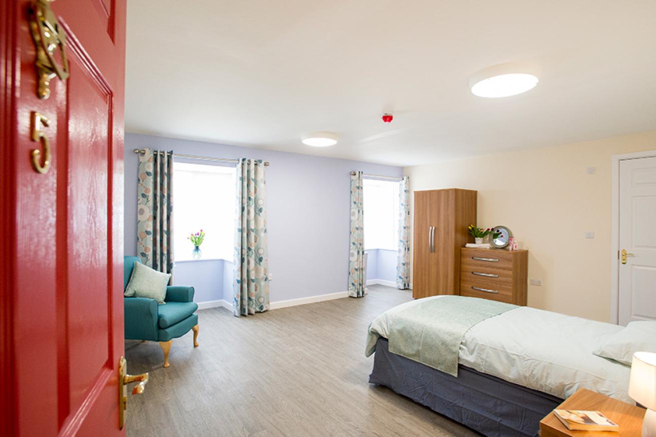 A large bedroom with natural light coming from two windows. There is a single bed, armchair, wardrobe and chest of draws in the room.