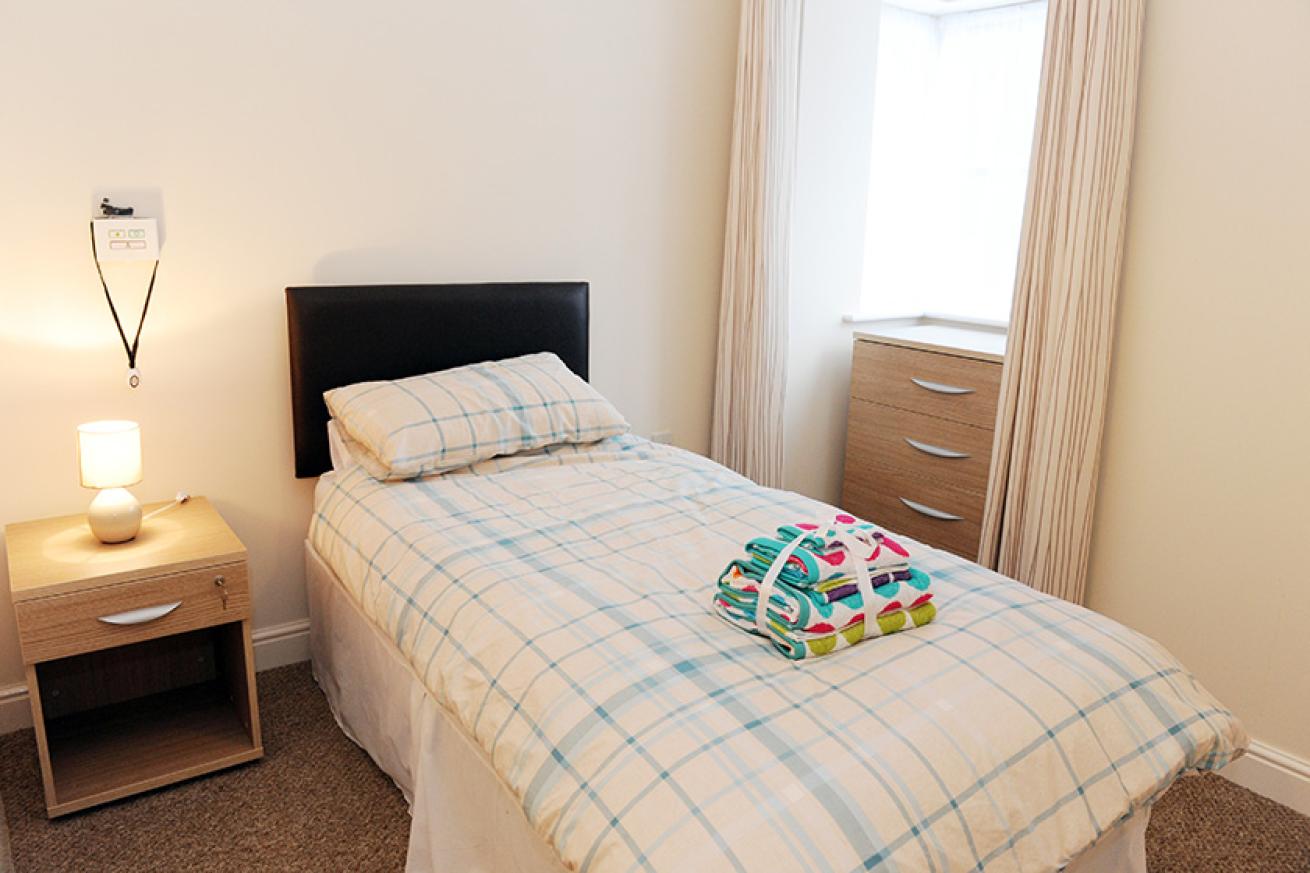 A small room painted cream which contains a single bed, bedside table with lamp, emergency pull cord and a chest of draws in front of a window where natural light is pouring in from