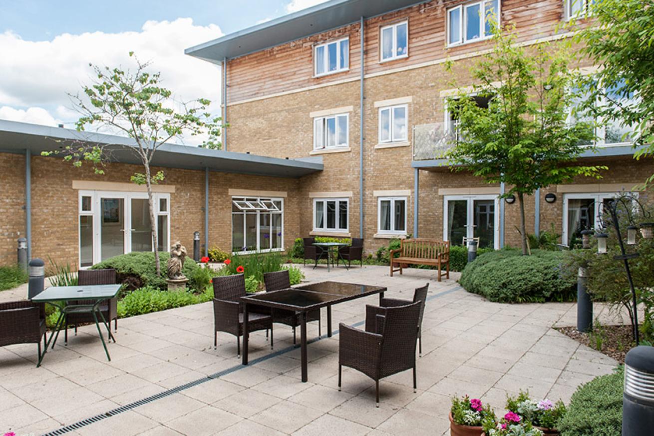 A patio garden with rattan furniture amongst flower beds and trees with the backdrop of a large retirement service building
