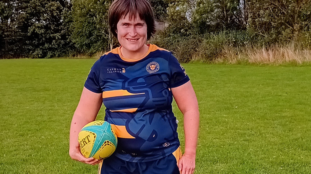 A woman wearing a rugby kit, holding a rugby ball and smiling at the camera.