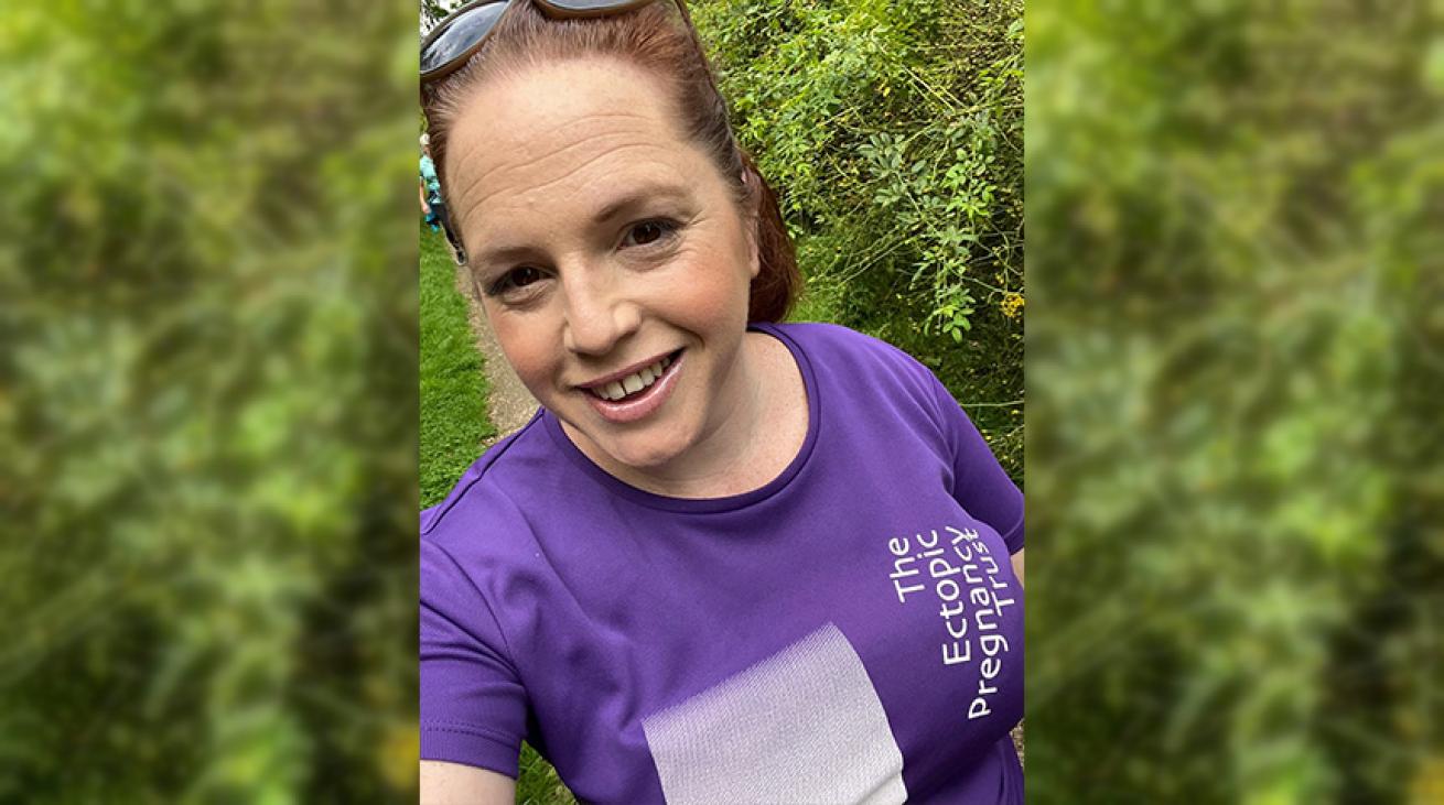 A woman wearing a purple t-shirt, walking and smiling while taking a selfie.