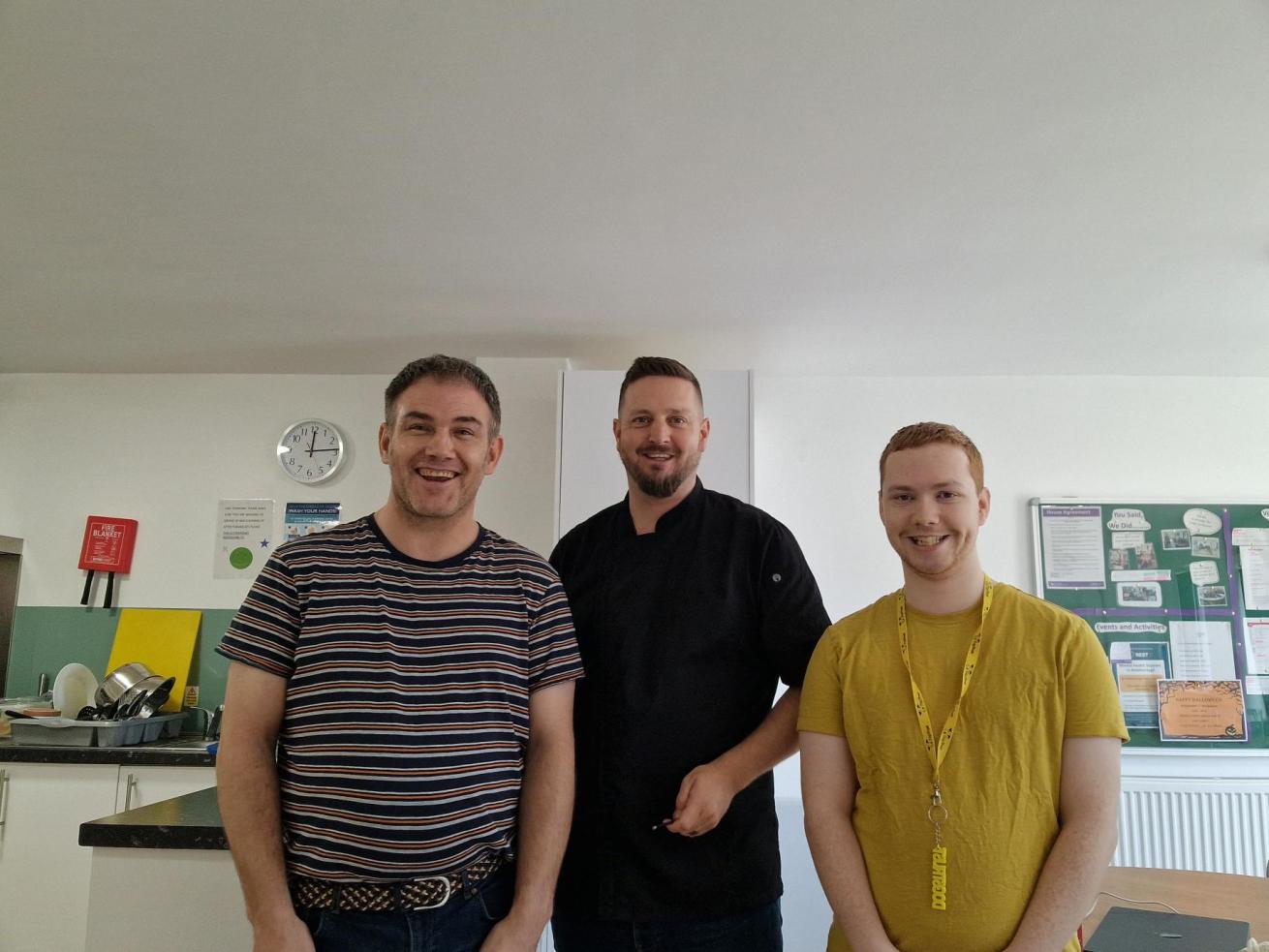 Three men stood in a kitchen and smiling at the camera, one wearing a chef's uniform.