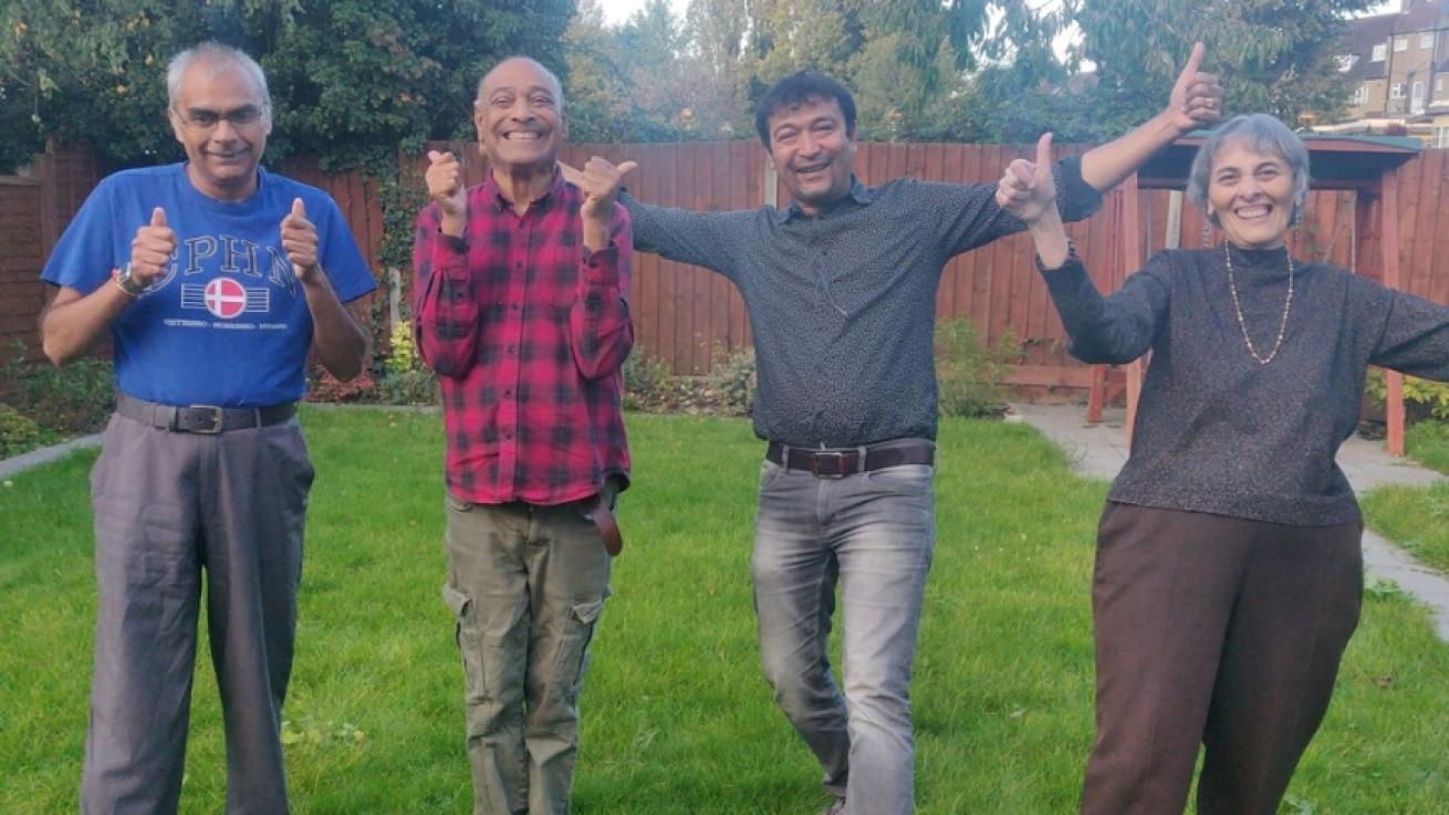 Three men and one woman standing in a garden, all smiling, giving two thumbs up and cheering to the camera.