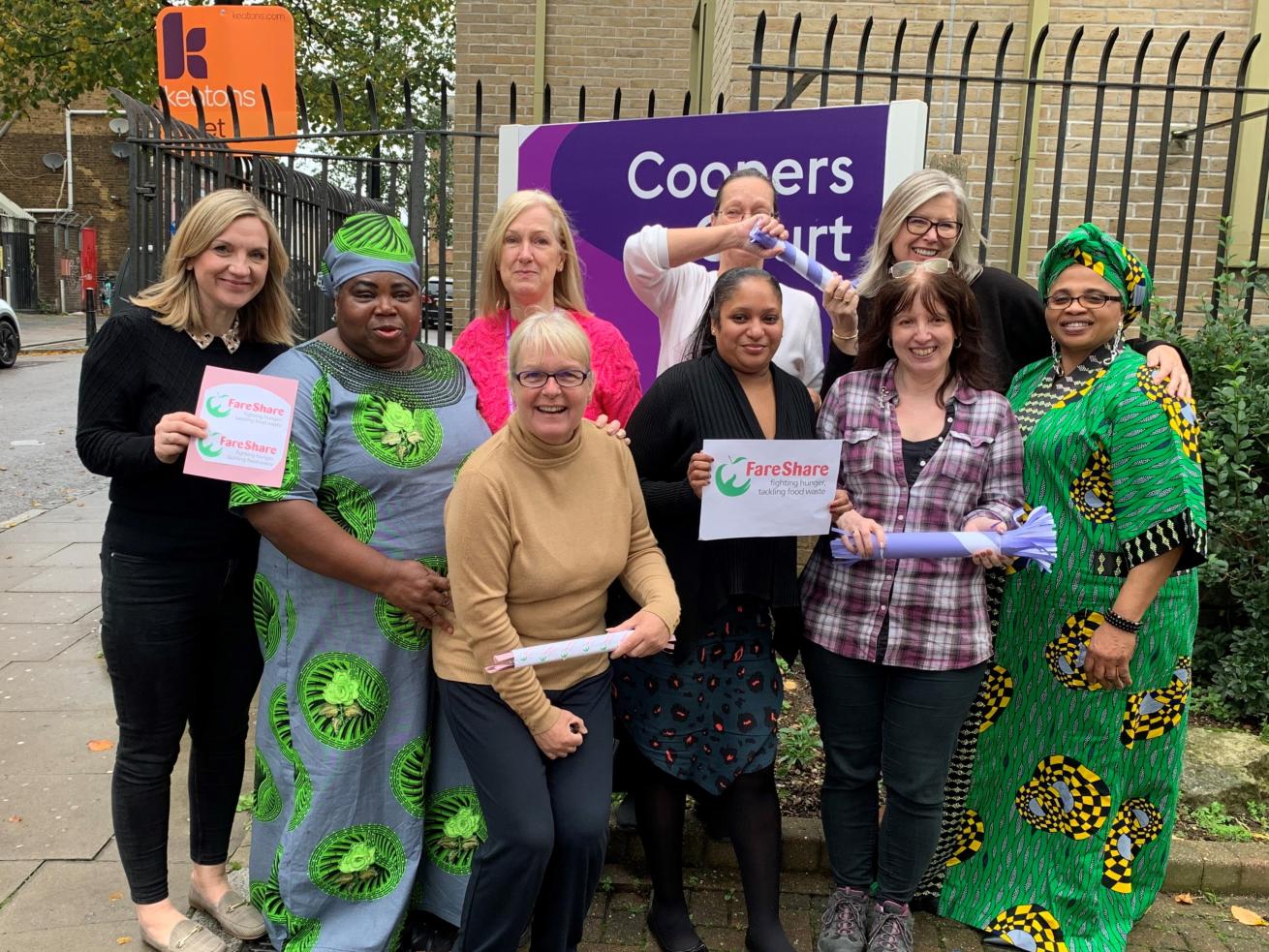 A group of nine women standing together in the street by a large sign that reads 'Coopers Court', smiling at the camera and holding signs that read 'FareShare'.