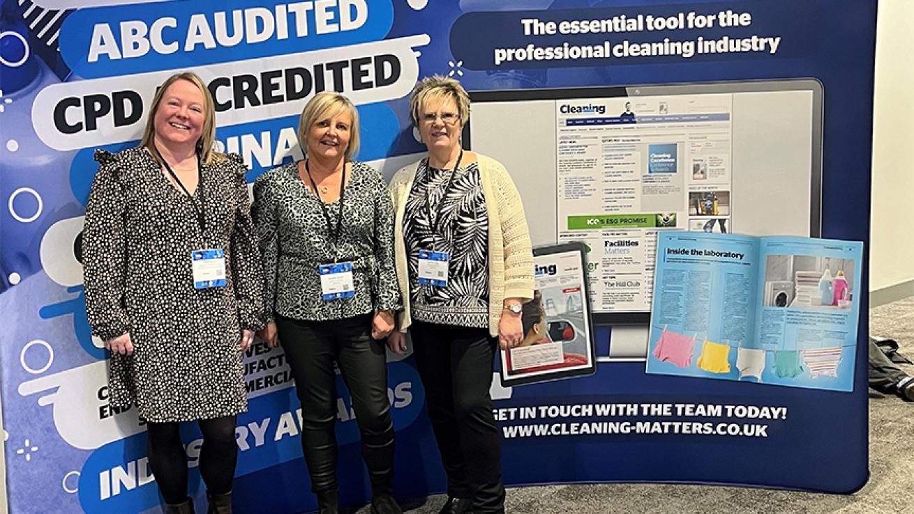 Three women wearing patterned tops and lanyards pose for a photograph in front of a board that reads "Cleaning matters"