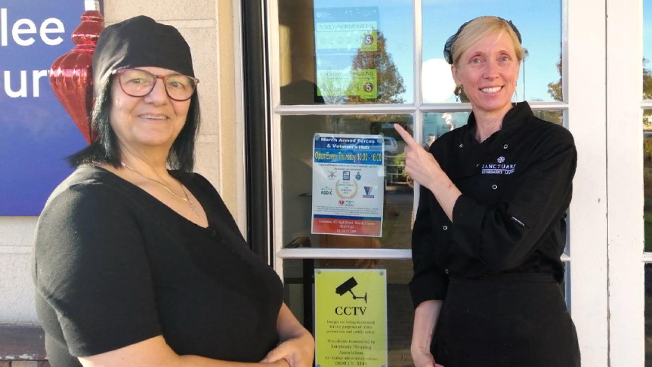 Two women smiling at the camera, pointing to a sign on a door that shows a rating of 5 from the Food Standards Agency.