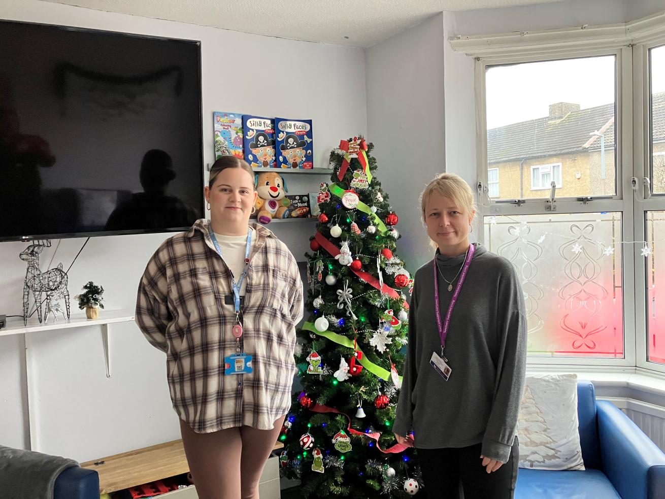 Two individuals standing next to a decorated Christmas tree in an indoor setting with children's books mounted on the wall behind them and a window letting in natural light