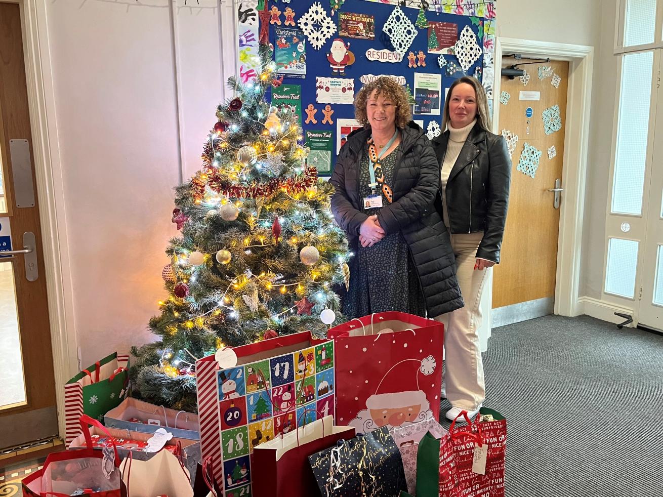 Two women stood by a Christmas tree and smiling at the camera, surrounded by gift bags containing presents.