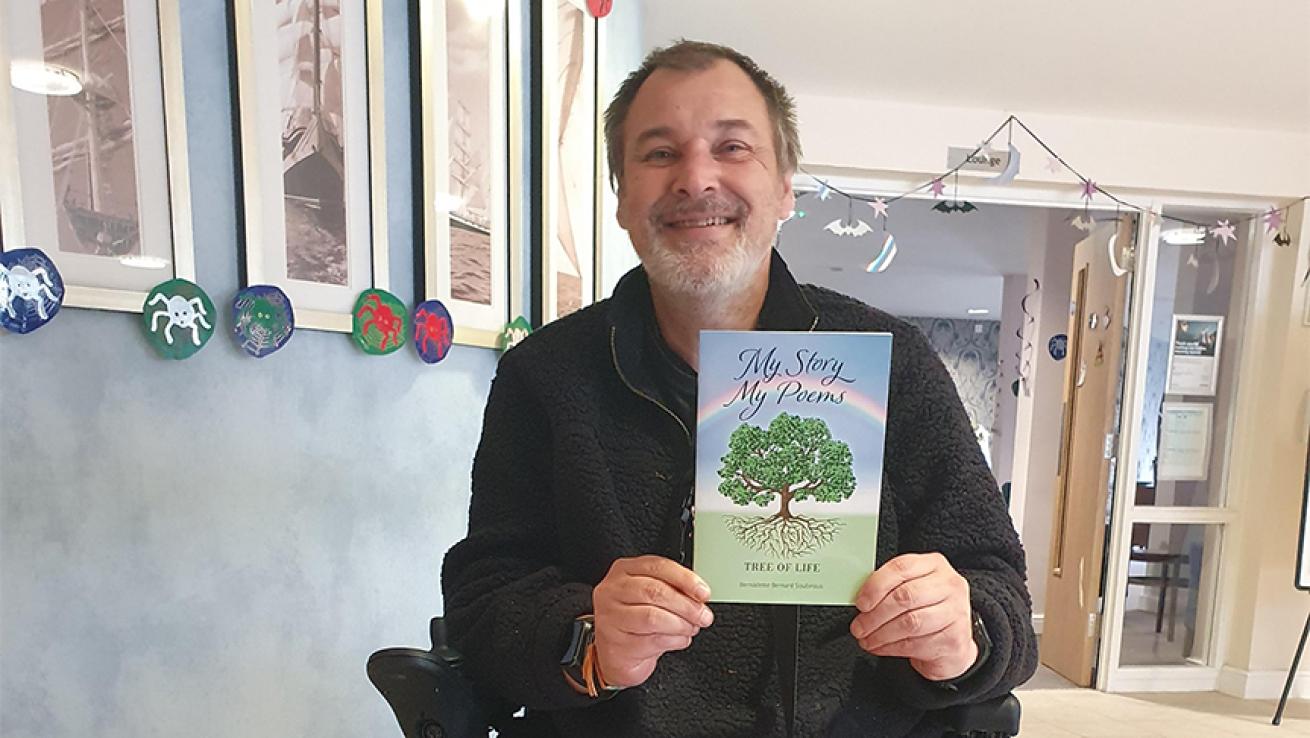 A bearded man proudly displays his poetry book titled "My Story, My Poems, Tree of Life." There is a rainbow on the front cover with a tree underneath