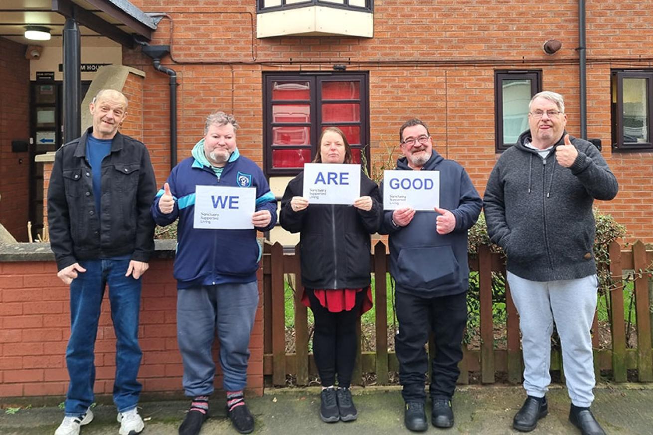 Five people standing in a row outside a building, smiling at the camera and holding signs saying 'We Are Good