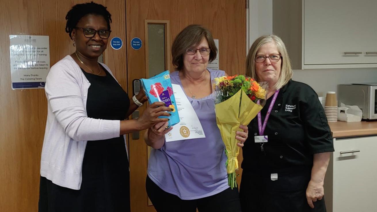Sanctuary chef pictured in a purple top, holding a certificate, a box of Roses chocolates and a bouquet of flowers. She stands between two colleagues in an indoor setting.