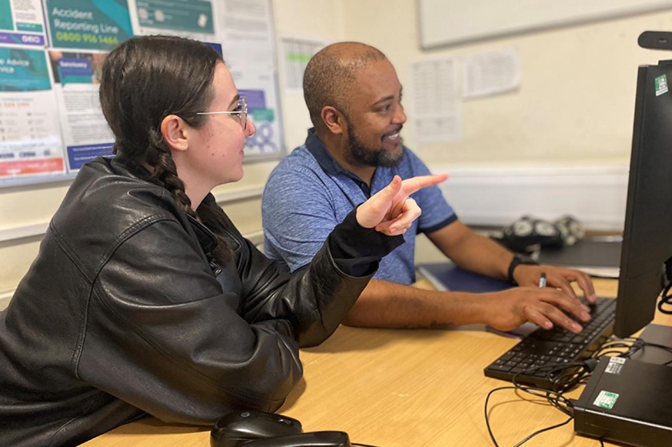 A woman and a man sat at a desk, looking at a computer together.