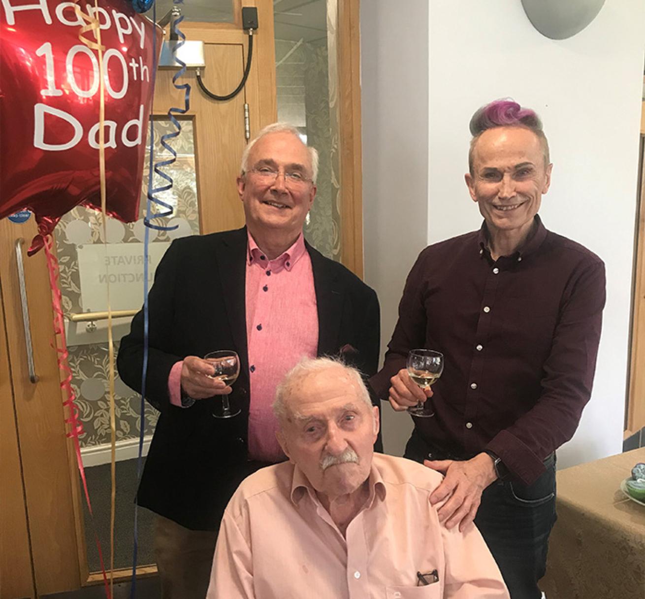 Three smartly dressed gentlemen pose for a photograph next to a red, star-shaped balloon that reads "Happy 100th Dad." The gentleman at the front is sitting in a wheelchair. He is elderly and wears a pastel pink shirt. The gentleman on the left behind him wears a salmon pink shirt and glasses and the gentleman on the right wears a purple mauve shirt, with a streak of purple in his hair to match. They both smile broadly with wine glasses in their hands.