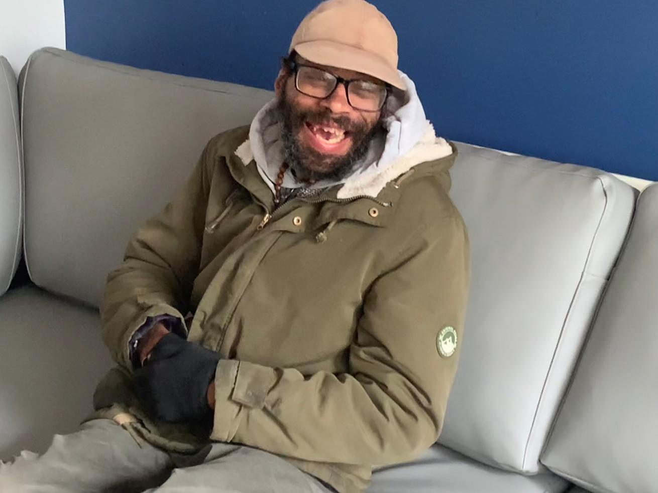 A black man wearing a green coat and hat, sat on a grey sofa against a blue wall, smiling at the camera