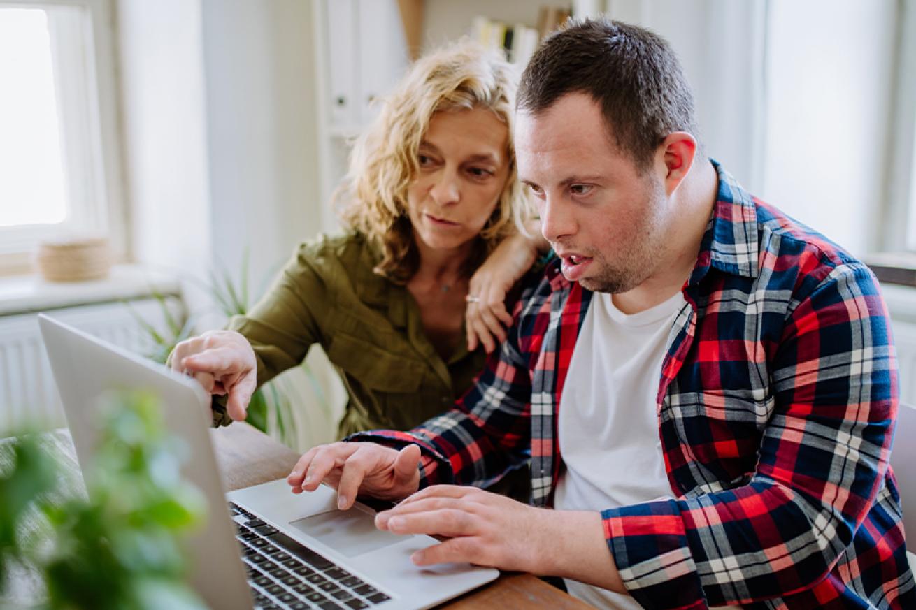 Stock photo of a young man with down syndrome wearing a red chequered shirt using a laptop on a wooden table with the help of an older woman wearing a green shirt