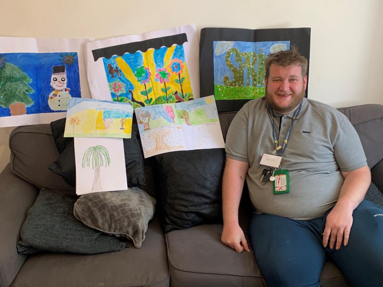 A white man with fair hair in his 30s, sat on a sofa in a lounge, surrounded by his hand painted artwork, smiling at the camera.
