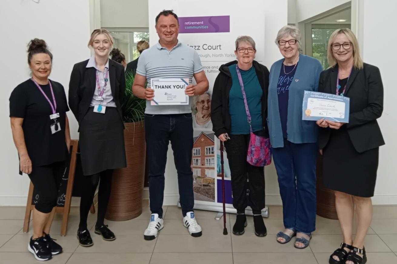 Five women standing in a row with one man in the centre, smiling at the camera and holding a cheque.