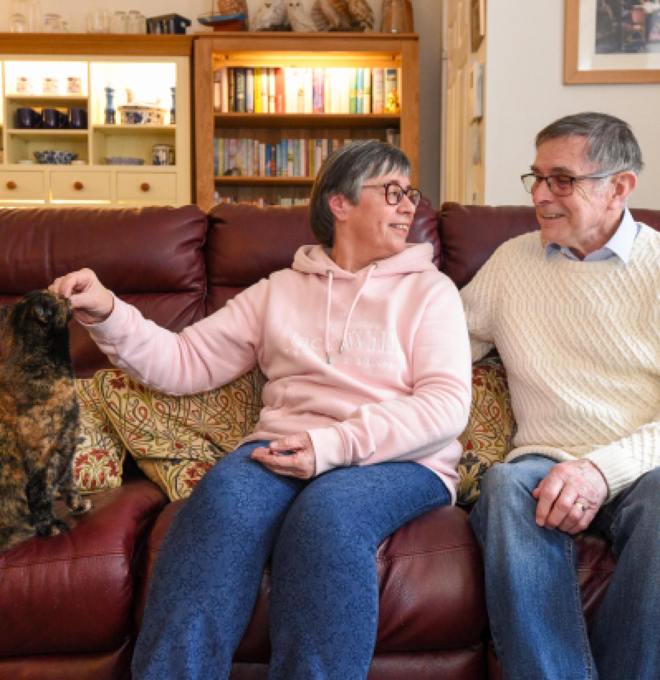 An elderly woman and man sitting on a burgundy leather sofa in a family living room. The woman is feeding an orange and black cat