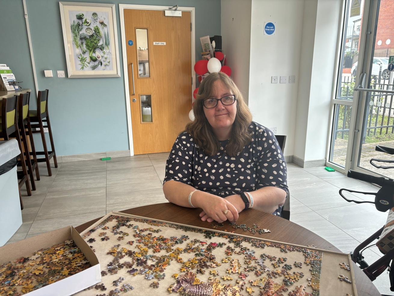 A white woman with shoulder-length brown hair and glasses, sat at a round table in front of a half-completed jigsaw showing giraffes, and smiling at the camera.