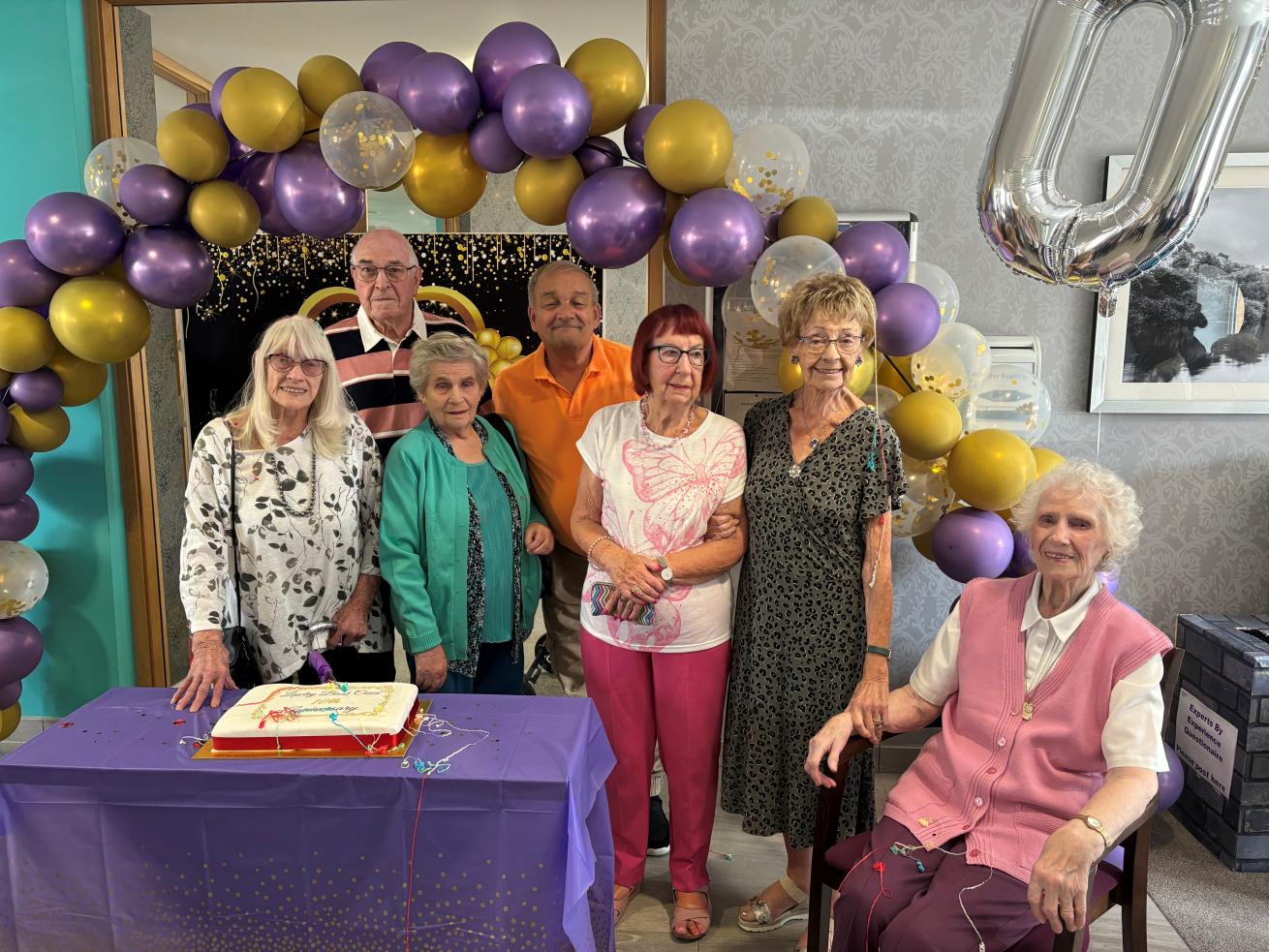 A group of people in their 70s and 80s smiling at the camera, surrounded by a purple and gold balloon arch, with a cake in front of them.