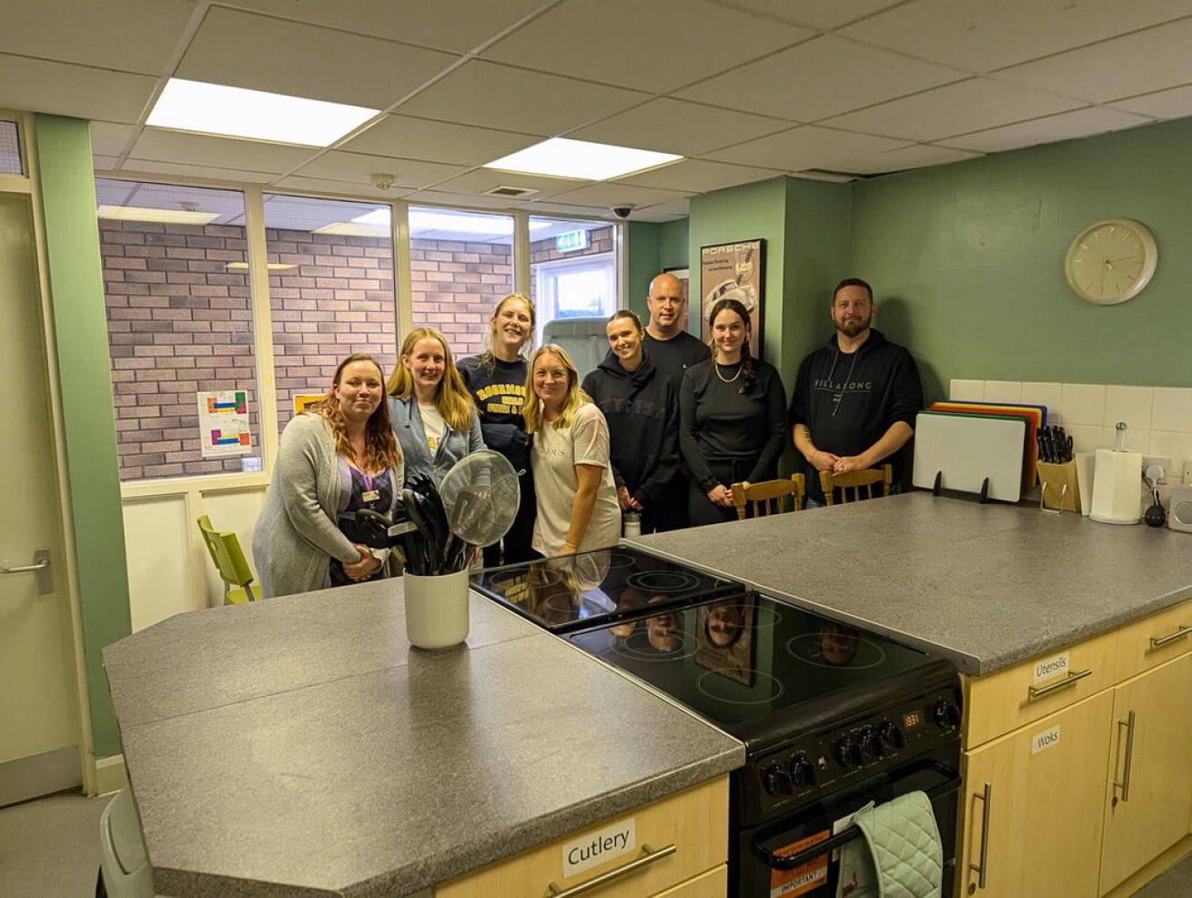  A group of eight white men and women in their 20s, 30s and 40s, standing together and smiling at the camera. They're stood in a kitchen with freshly-painted green walls. In front of them is a kitchen island with two cookers and a pot of utensils. They're all smiling at the camera.