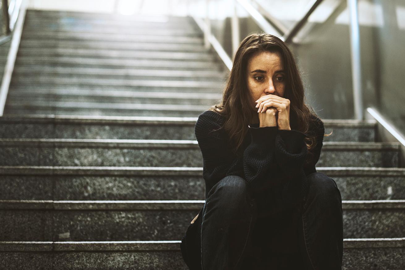 A woman in her 20s wearing dark clothes and with long dark hair, sat on the dark stairs like those which lead down to an underground station or walkway. She looks worried and has her hands clenched in front of her face.