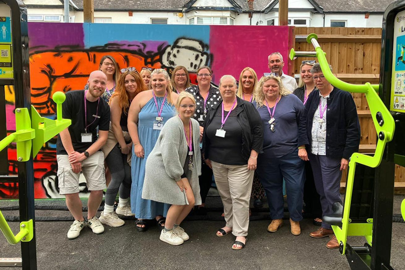 A group of male and female Sanctuary Supported Living staff stand in front of a graffiti mural, smiling. Most wear purple lanyards, and lime green gym equipment like a Lat Press is visible in the foreground.