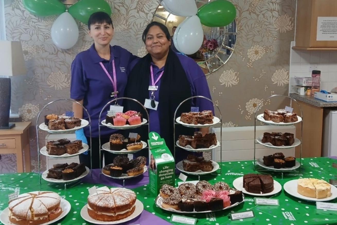 Two women wearing purple Sanctuary Supported Living branded shirts standing behind a table covered in cakes and pastries on stands and plates