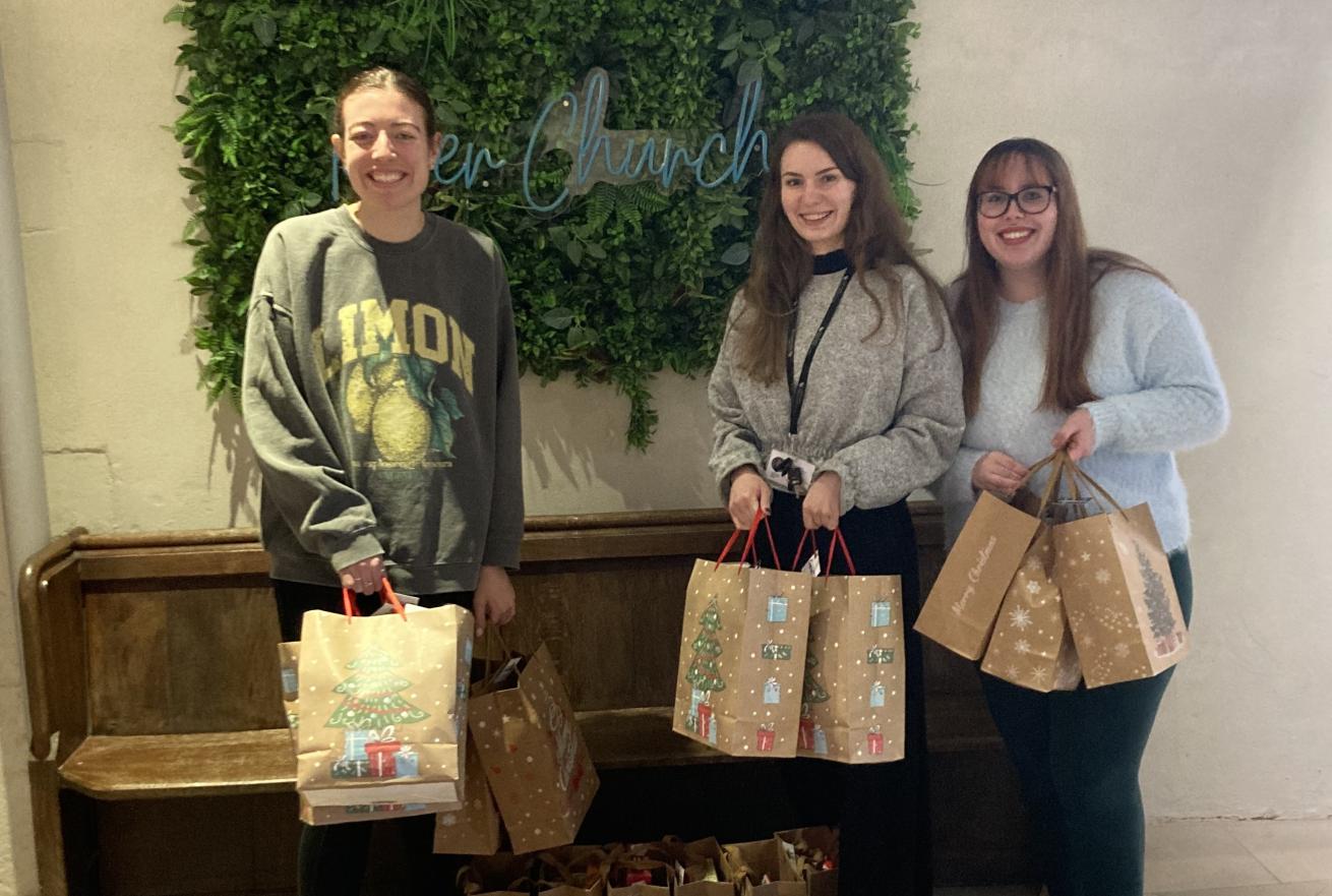 Three white women in their 30s standing inside the entrance to a pale-walled modern church, with a 'River Church' sign behind them. They're smiling at the camera and surrounded by brown paper Christmas gift bags laid out across the floor.