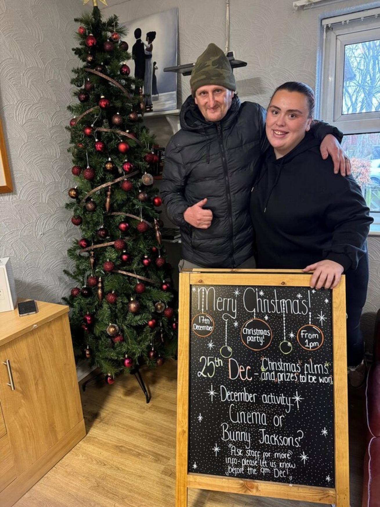 A man in a green camo beanie and black quilted coat gives a thumbs-up while standing next to a woman in a black hoody. They stand behind a blackboard with Christmas activity details, near a Christmas tree decorated with red baubles.