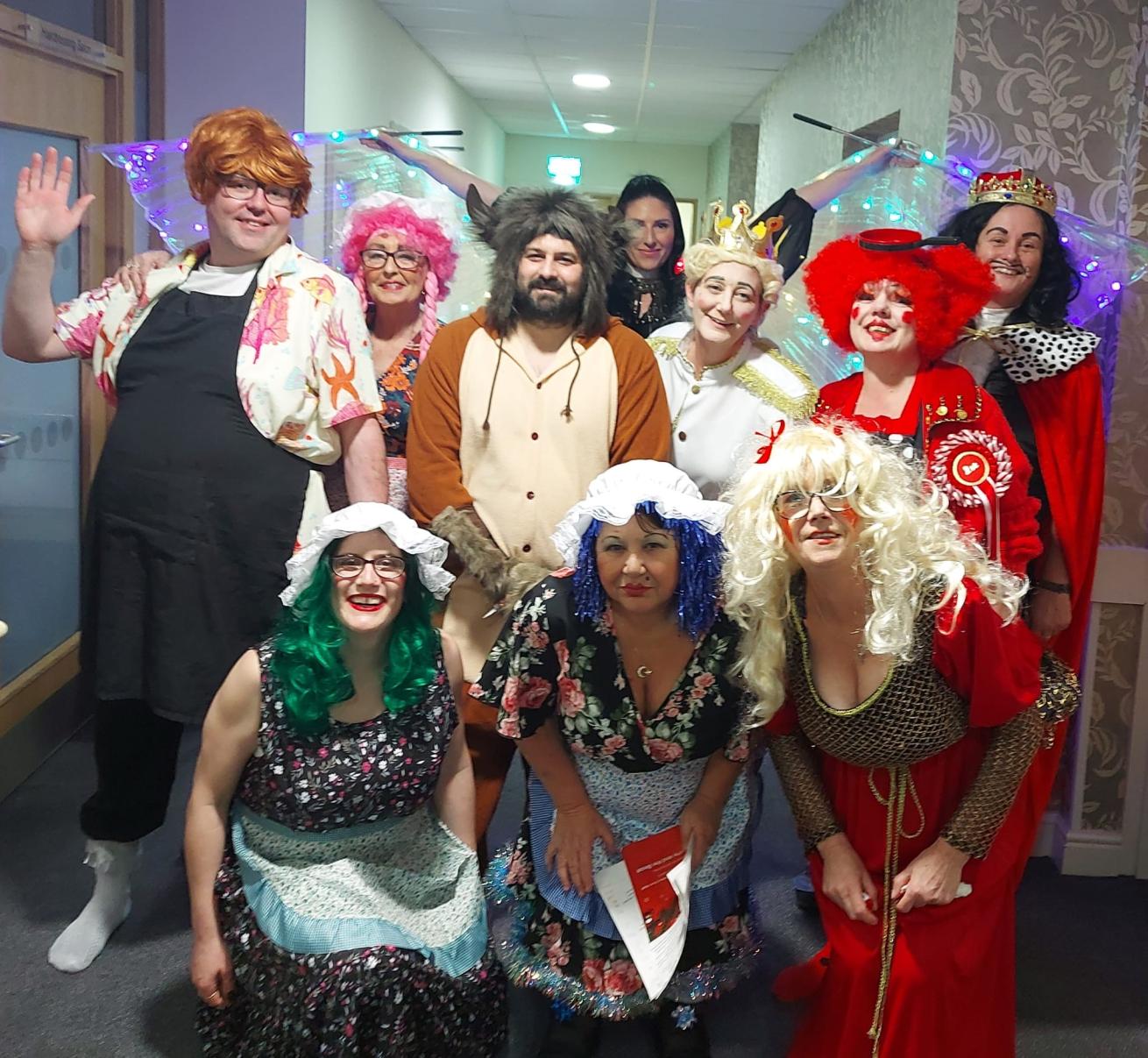A group of 10 staff members from Whitley Court and Dunboyne Court dressed as characters from the pantomime 'Smoochy and the Beast,' wearing vibrant costumes, bold makeup, and colourful, extravagant wigs.
