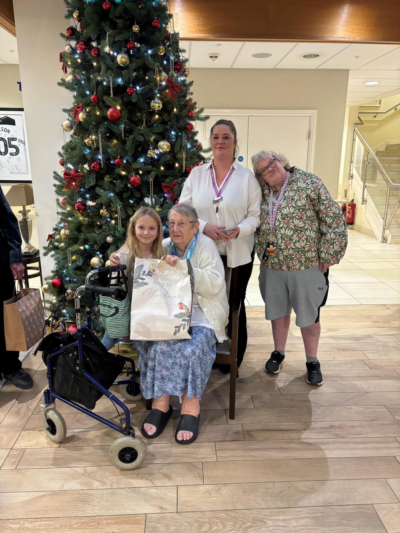 An older lady wearing a blue dress and white cardigan seated by a large Christmas tree in the bright atrium of a retirement community, with a young blond-haired girl next to her, holding a large Christmas gift bag. Behind them is a lady in her 30s wearing a white blouse and purple lanyard, with a lady in her late 50s or 60s stood next to her. All are smiling at the camera.