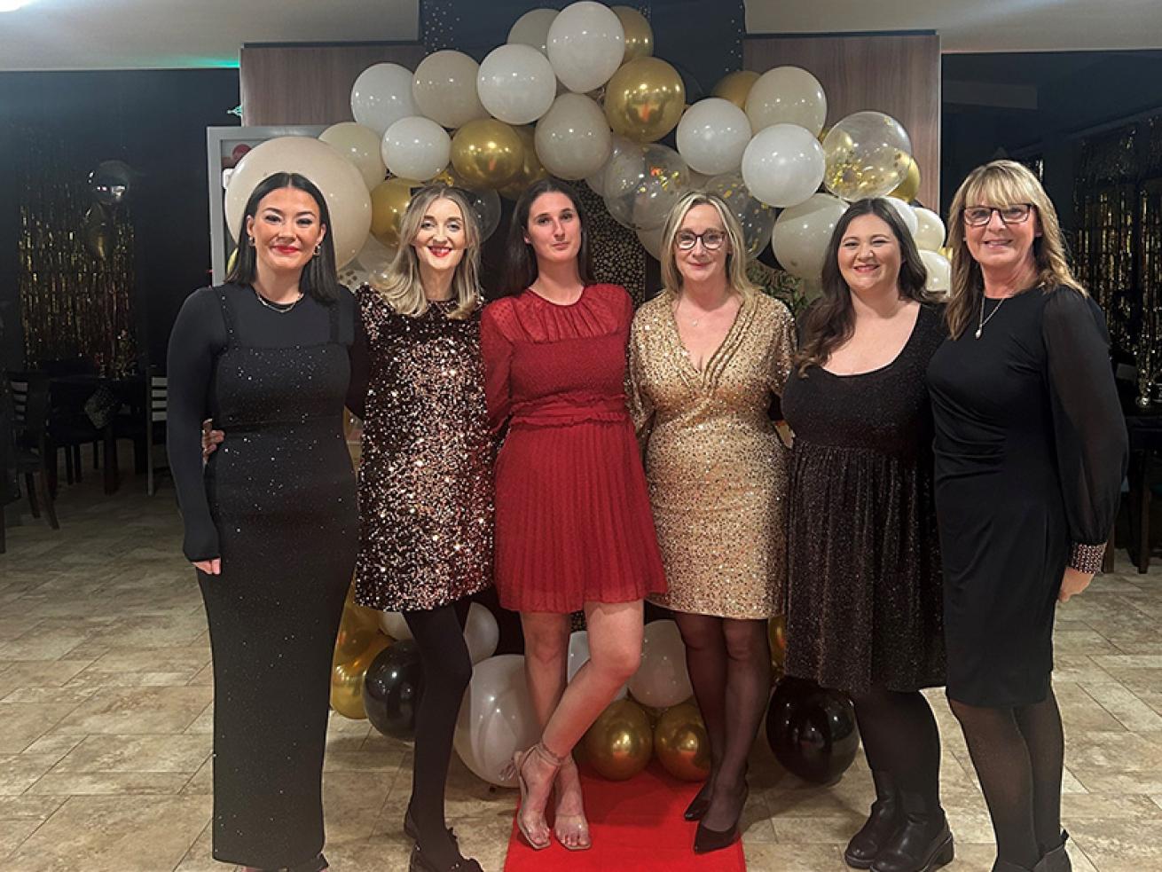 Six white women aged from their 20s to their 50s, all dressed in black, red or gold party dresses, stood inside a retirement community large communal room, on top of a red carpet leading to a white and gold balloon arch . They're stood together smiling at the camera.