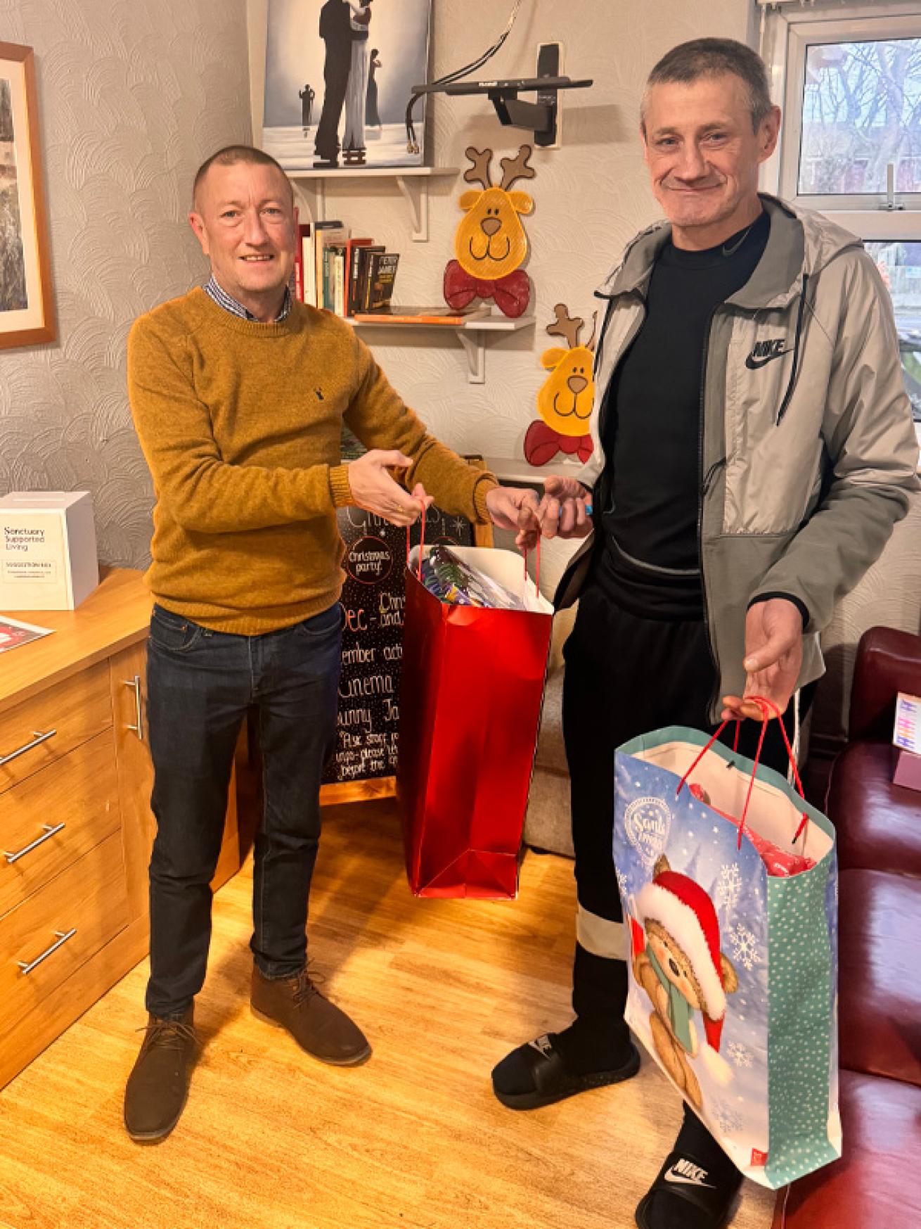 Two men standing together, the first wearing a mustard coloured jumper and dark wash jeans, and the second, taller man in a Nike tracksuit top. They are holding Christmas gift bags and smiling.