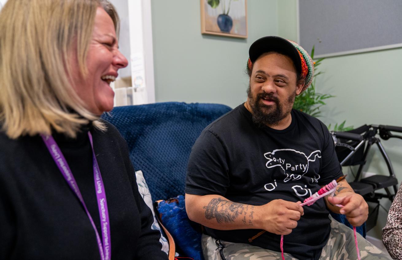 A white woman with blonde hair wearing a purple Sanctuary Supported Living lanyard laughs next to a man with darker skin, wearing a rastacap, sideburns, and a beard, using crochet needles with pink and white wool
