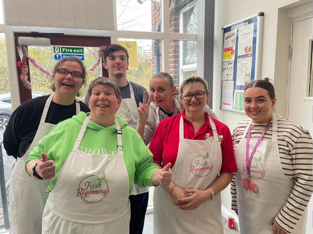 A group of six people standing together indoors, all wearing white aprons with the "Fresh Beginnings" logo. They are smiling and posing for the camera. One person is giving a thumbs-up, while another is making a peace sign. They appear to be in a kitchen or community space.